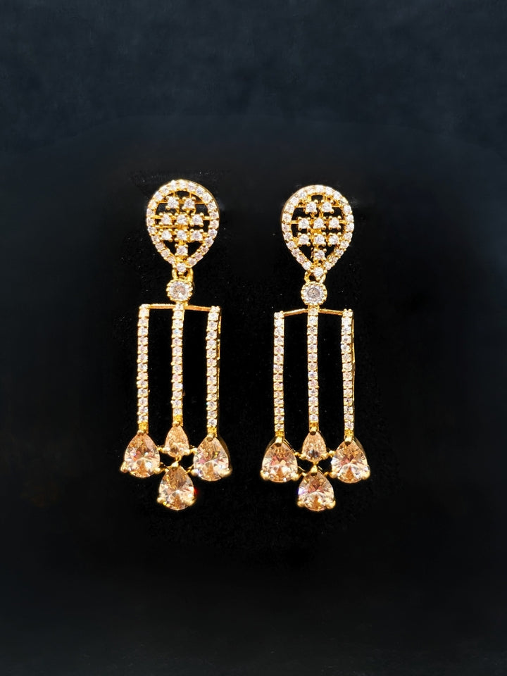 The Lavanya Necklace Set features a gold Champaign necklace with vertical bars topped by teardrop-shaped champagne colored American Diamonds, paired with matching dangling earrings and displayed on a blue velvet bust with ornate embroidery on the left.