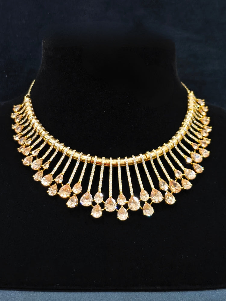 The Lavanya Necklace Set features a gold Champaign necklace with vertical bars topped by teardrop-shaped champagne colored American Diamonds, paired with matching dangling earrings and displayed on a blue velvet bust with ornate embroidery on the left.