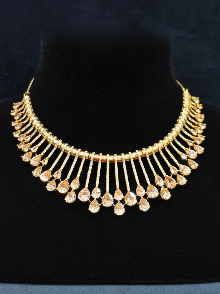 The Lavanya Necklace Set features a gold Champaign necklace with vertical bars topped by teardrop-shaped champagne colored American Diamonds, paired with matching dangling earrings and displayed on a blue velvet bust with ornate embroidery on the left.