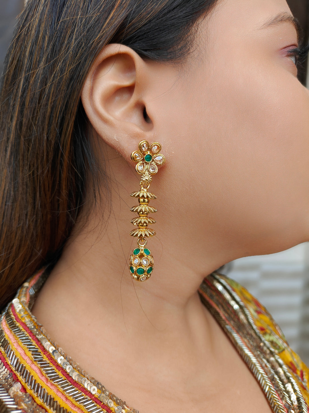 A close-up of a womans ear wearing Laxmi Green Earrings featuring a floral design with dangling gold elements and green gemstone accents at the oval drop at the bottom —ideal for wedding ceremony jewelry.