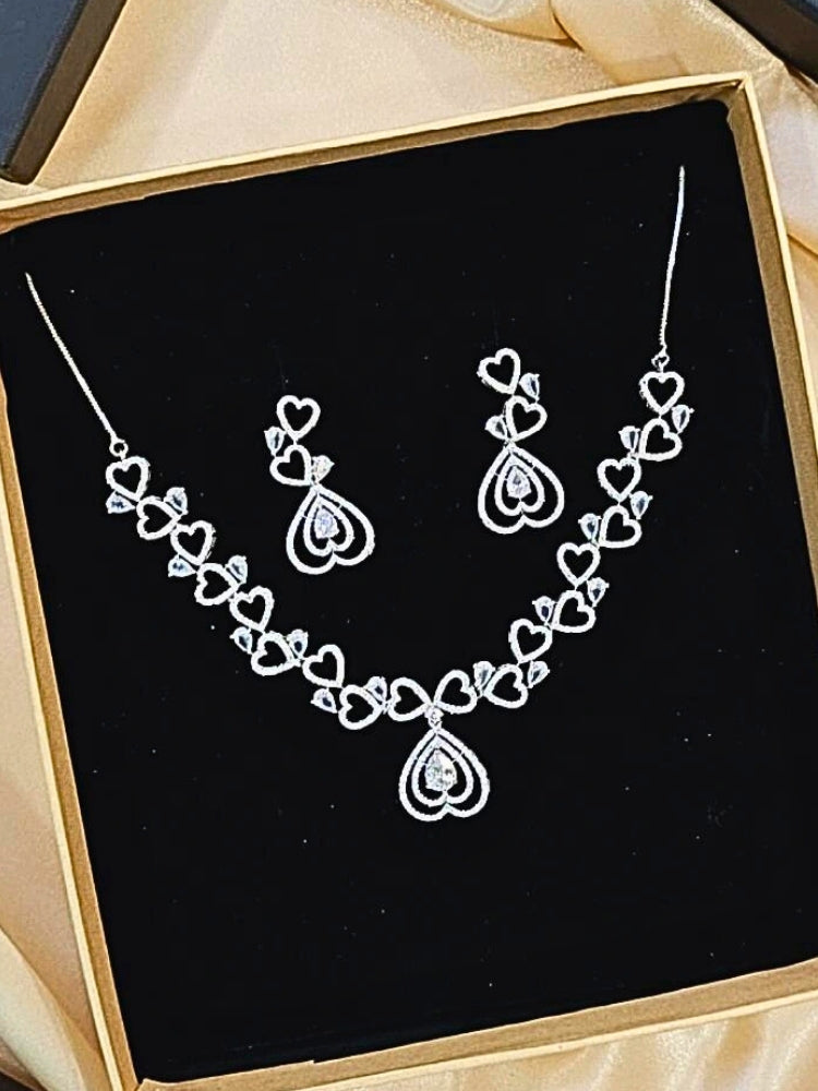 The Love Necklace Set features a heart-shaped white necklace and matching earrings with sparkling American Diamonds, elegantly presented in a black velvet box—perfect to complement your favorite cocktail gown.