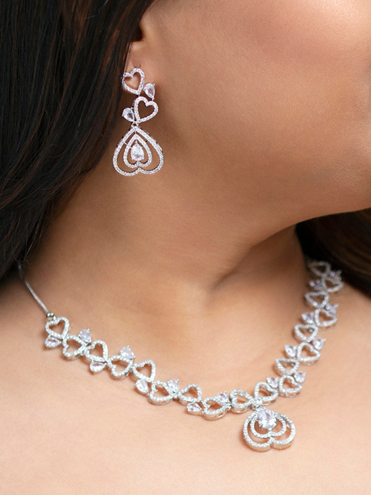 The Love Necklace Set features a heart-shaped white necklace and matching earrings with sparkling American Diamonds, elegantly presented in a black velvet box—perfect to complement your favorite cocktail gown.