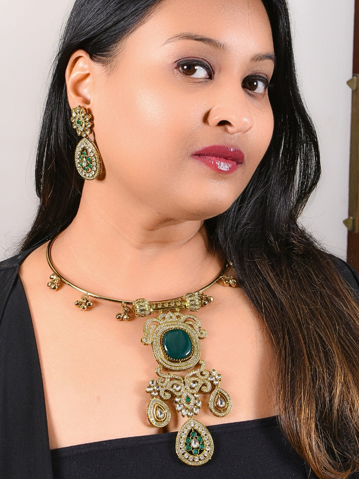 A woman with long dark hair wears a black top and the Lupin Dark Green Necklace Set, featuring ornate ethnic jewelry with emerald green accents and matching earrings. The hasli necklace set is made in mehendi polish, along with emerald green stones and american diamonds. 