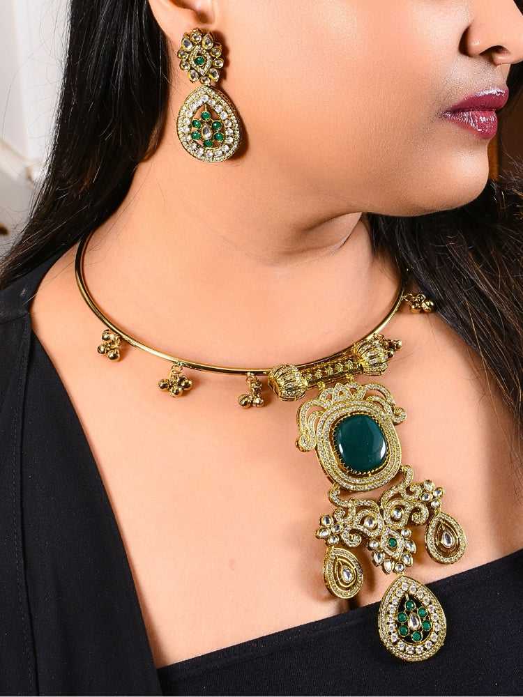 A woman with long dark hair wears a black top and the Lupin Dark Green Necklace Set, featuring ornate ethnic jewelry with emerald green accents and matching earrings. The hasli necklace set is made in mehendi polish, along with emerald green stones and american diamonds. 