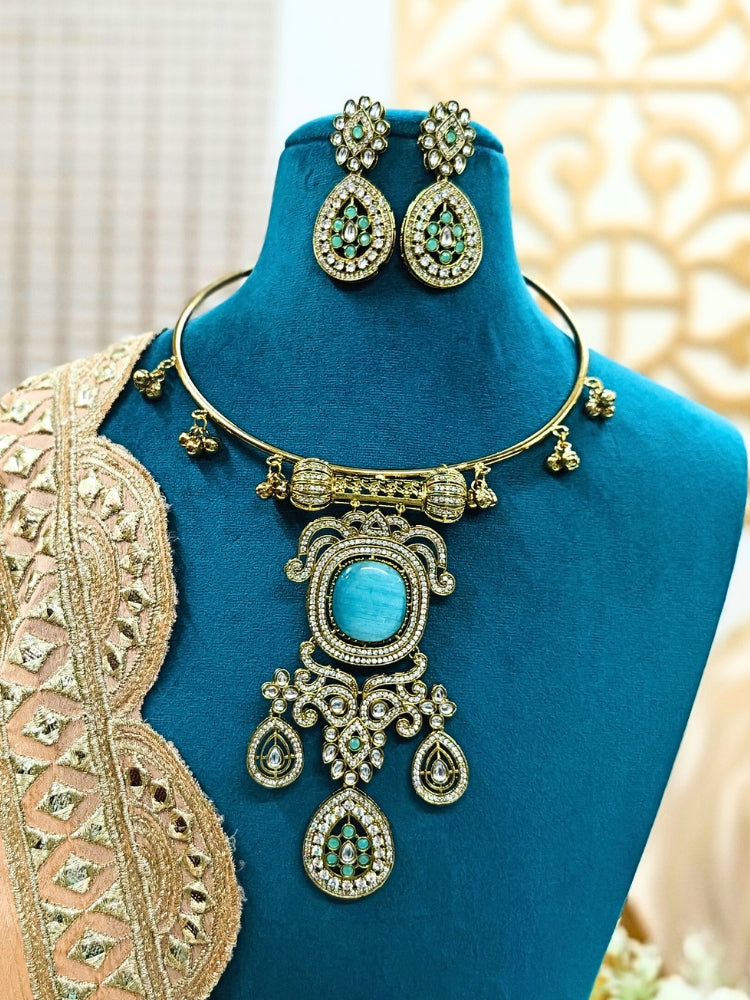 Discover the elegance of the Lupin Hasli Necklace Set, featuring a radiant Mint Blue Monalisa stone, sparkling American diamonds, and charming tiny ghungroos. Perfect for saris, weddings, and festive occasions. Put on a blue mannequin, and a beige embroidered fabric draped over the shoulder