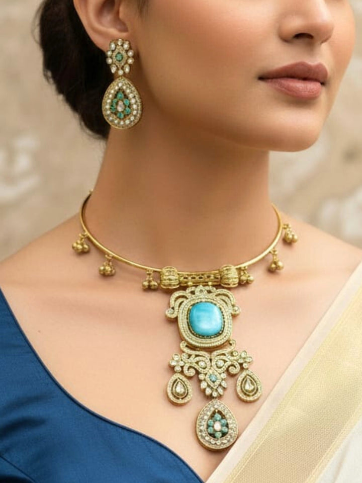 Discover the elegance of the Lupin Hasli Necklace Set, featuring a radiant Mint Blue Monalisa stone, sparkling American diamonds, and charming tiny ghungroos. Perfect for saris, weddings, and festive occasions. Put on a blue mannequin, and a beige embroidered fabric draped over the shoulder