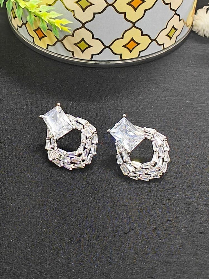 The Mabel White Earrings feature sparkling American Diamonds with square-cut stones on top and circular woven baguette designs below, shown on a black surface with a patterned container in the background.