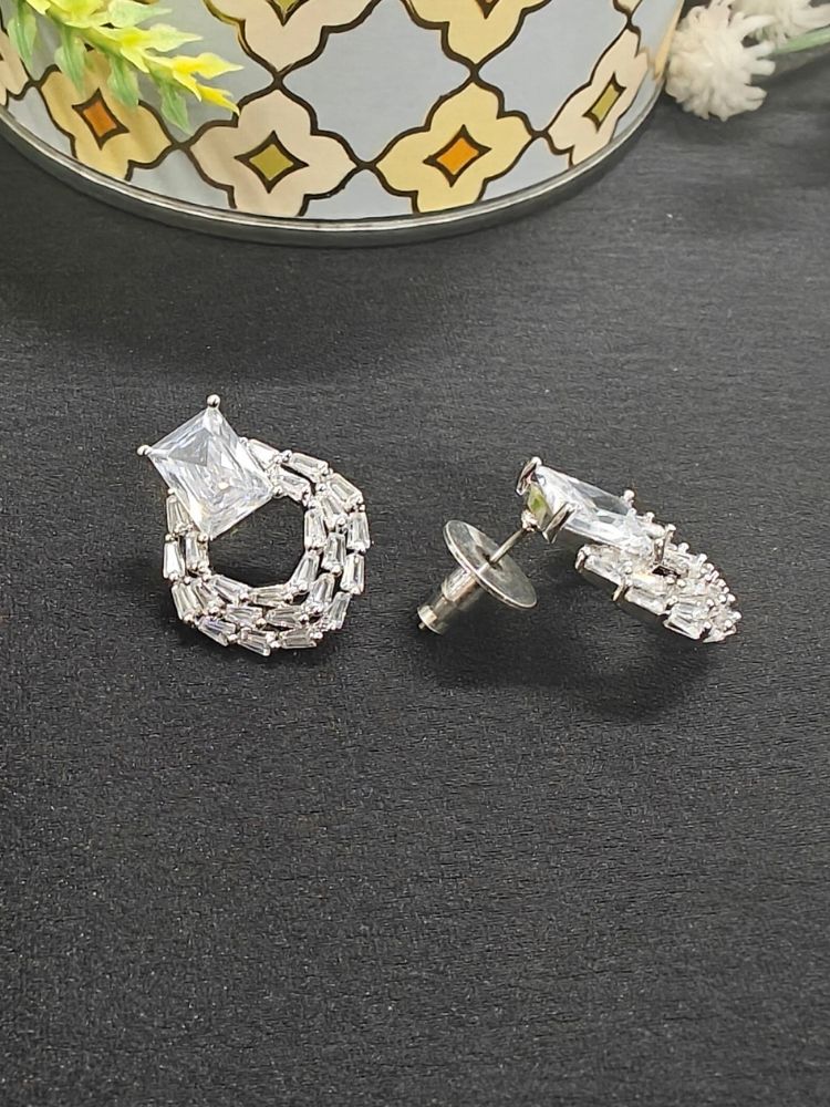 The Mabel White Earrings feature sparkling American Diamonds with square-cut stones on top and circular woven baguette designs below, shown on a black surface with a patterned container in the background.