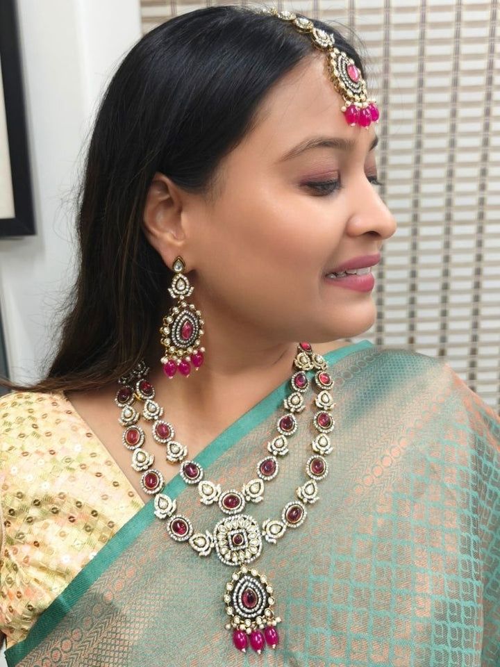 A woman in a green and gold saree, wearing the Magenta Pink Shabana Necklace Set, including an elaborate 2-layered necklace, earrings, and a mangtikka with pink colored stones and american diamonds, smiles warmly while looking to the side.