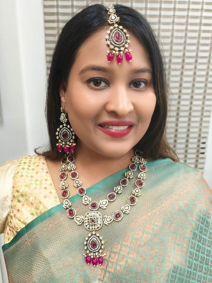A woman in a green and gold saree, wearing the Magenta Pink Shabana Necklace Set, including an elaborate 2-layered necklace, earrings, and a mangtikka with pink colored stones and american diamonds, smiles warmly while looking to the side.