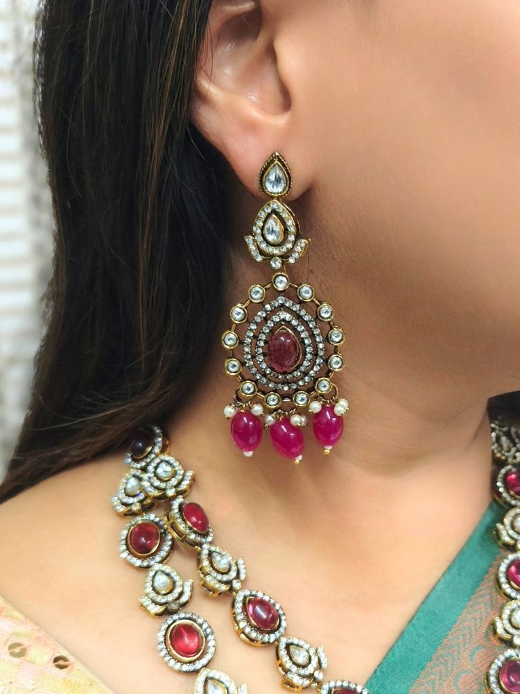 A woman in a green and gold saree, wearing the Magenta Pink Shabana Necklace Set, including an elaborate 2-layered necklace, earrings, and a mangtikka with pink colored stones and american diamonds, smiles warmly while looking to the side.