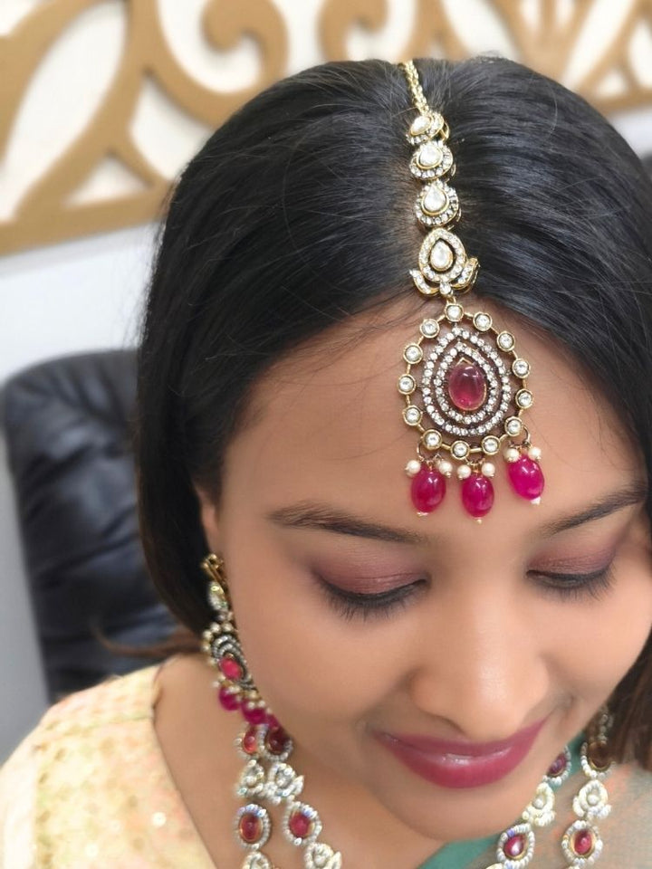 A woman in a green and gold saree, wearing the Magenta Pink Shabana Necklace Set, including an elaborate 2-layered necklace, earrings, and a mangtikka with pink colored stones and american diamonds, smiles warmly while looking to the side.