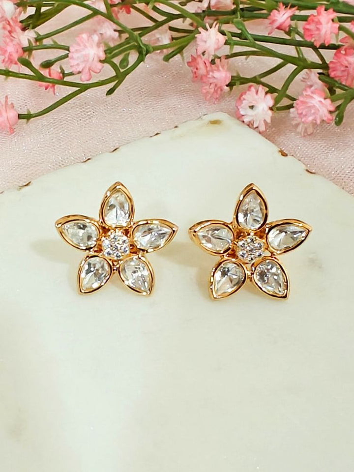 The Magnolia tyani kundan stud Earrings are gold, flower-shaped with clear teardrop American diamond petals and round diamonds at the center, shown on a white surface accented by pink flowers in the background.