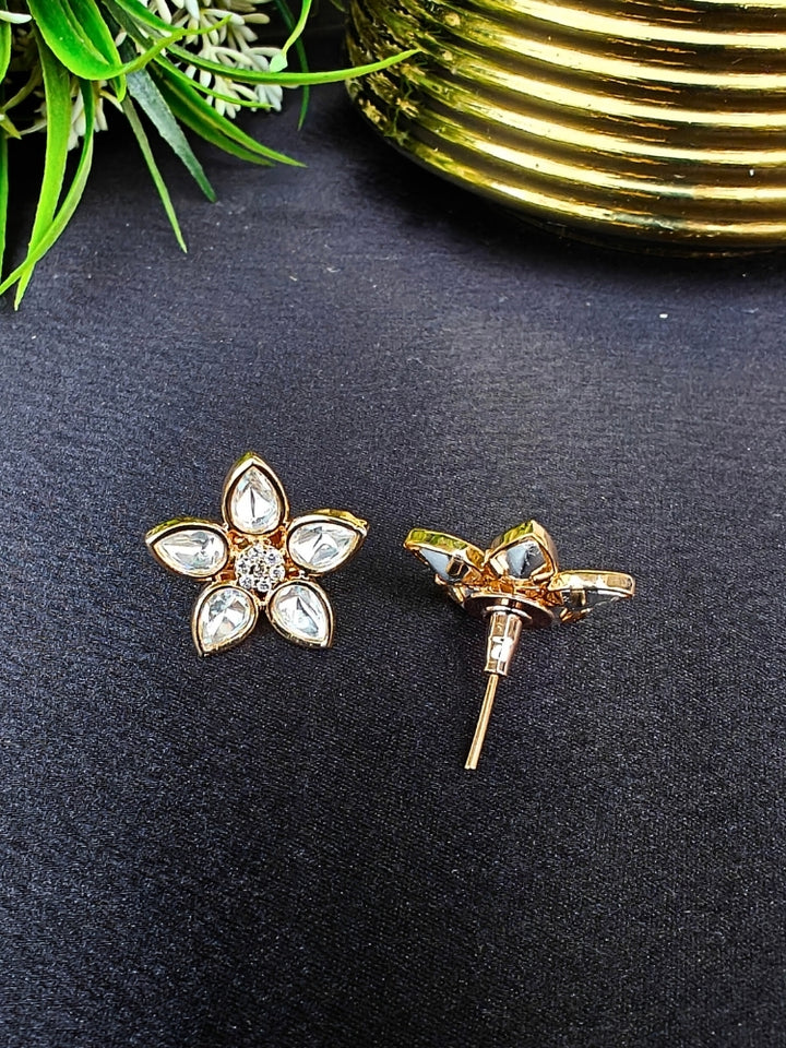 The Magnolia tyani kundan stud Earrings are gold, flower-shaped with clear teardrop American diamond petals and round diamonds at the center, shown on a Black surface accented by Leaves in the background.