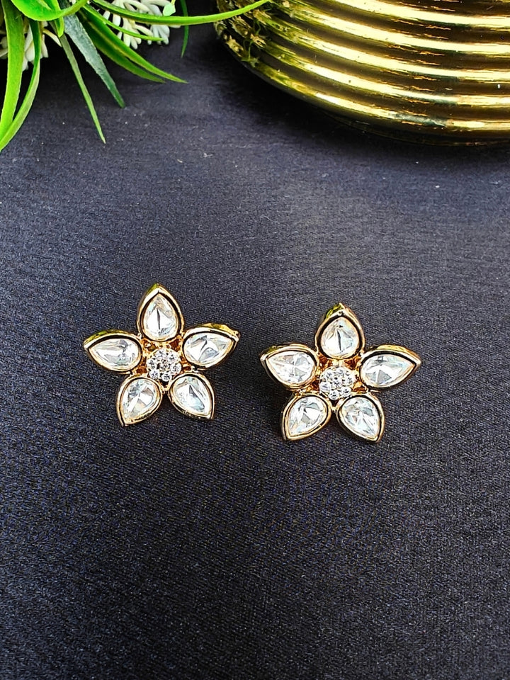 The Magnolia tyani kundan stud Earrings are gold, flower-shaped with clear teardrop American diamond petals and round diamonds at the center, shown on a Black surface accented by Leaves in the background.