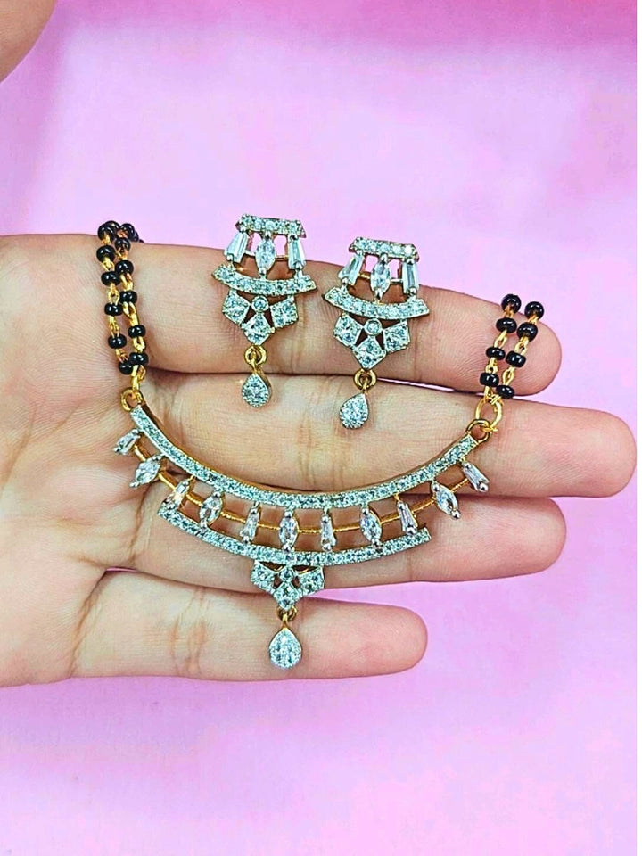 A hand holds the Mahira Mangalsutra—a gold necklace with black mangalsutra beads and matching earrings, pendant featuring sparkling American Diamonds—set against a soft pink background, perfect for your wedding ceremony dress. Diamond mangalsutra. Modern mangalsutra. 