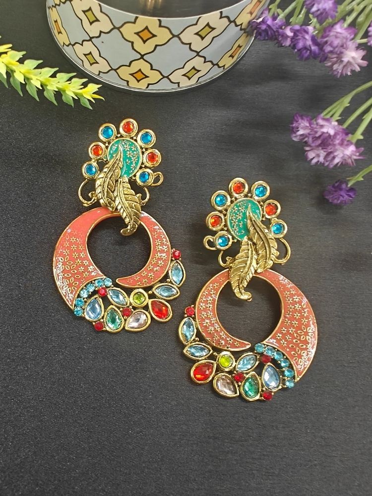 The Malika Multi-Color Earrings feature ornate Meenakari work with gold accents, circular shapes, leaf motifs, and red, blue, green, and clear stones. Displayed on black with purple and yellow flowers nearby.