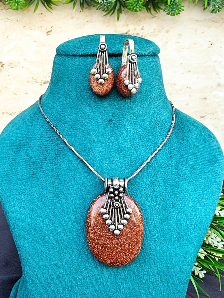 The Mandy Brown Pendant Set is displayed on a teal stand, featuring a necklace with a large Brown pendant and matching Brown oval earrings accented with colored stones, silver details, and small beads. Greenery decorates the background.
