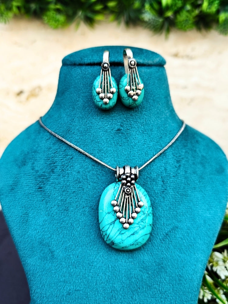 The Mandy Green Pendant Set is displayed on a teal stand, featuring a necklace with a large Green pendant and matching Green oval earrings accented with colored stones, silver details, and small beads. Greenery decorates the background.