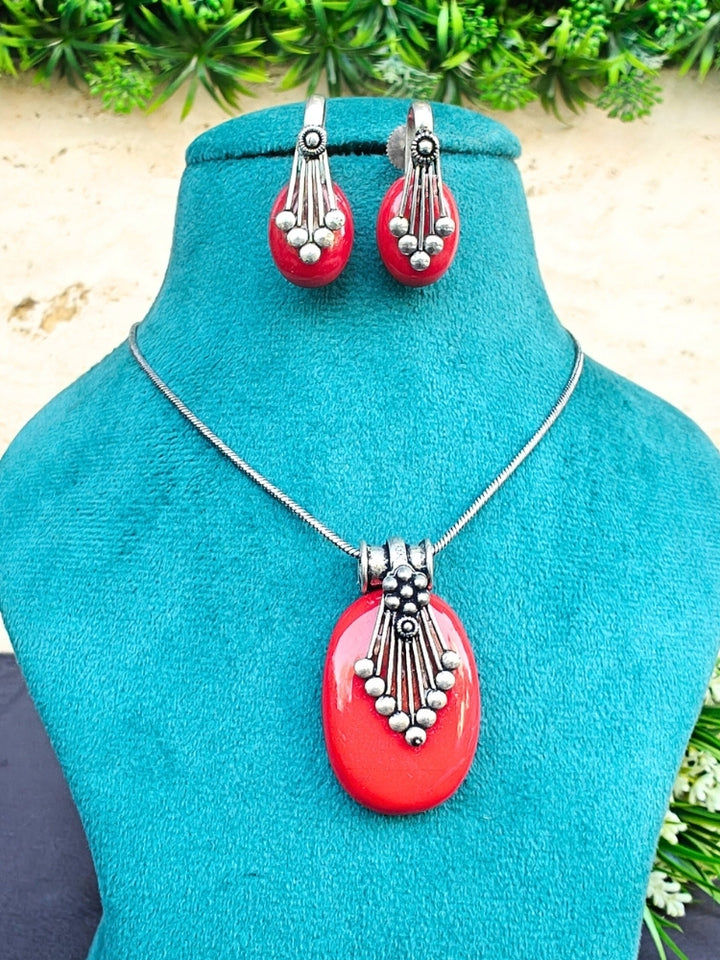 The Mandy Red Pendant Set is displayed on a teal stand, featuring a necklace with a large red pendant and matching red oval earrings accented with colored stones, silver details, and small beads. Greenery decorates the background.