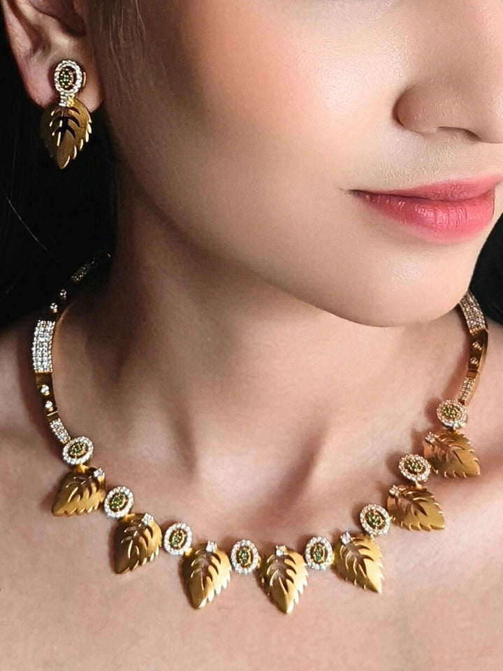 The Maple Green Necklace Set, featuring gold and green tones with matching leaf-shaped pendants and sparkling American Diamonds, is displayed on a black surface with flowers—ideal for wedding ceremony jewelry.