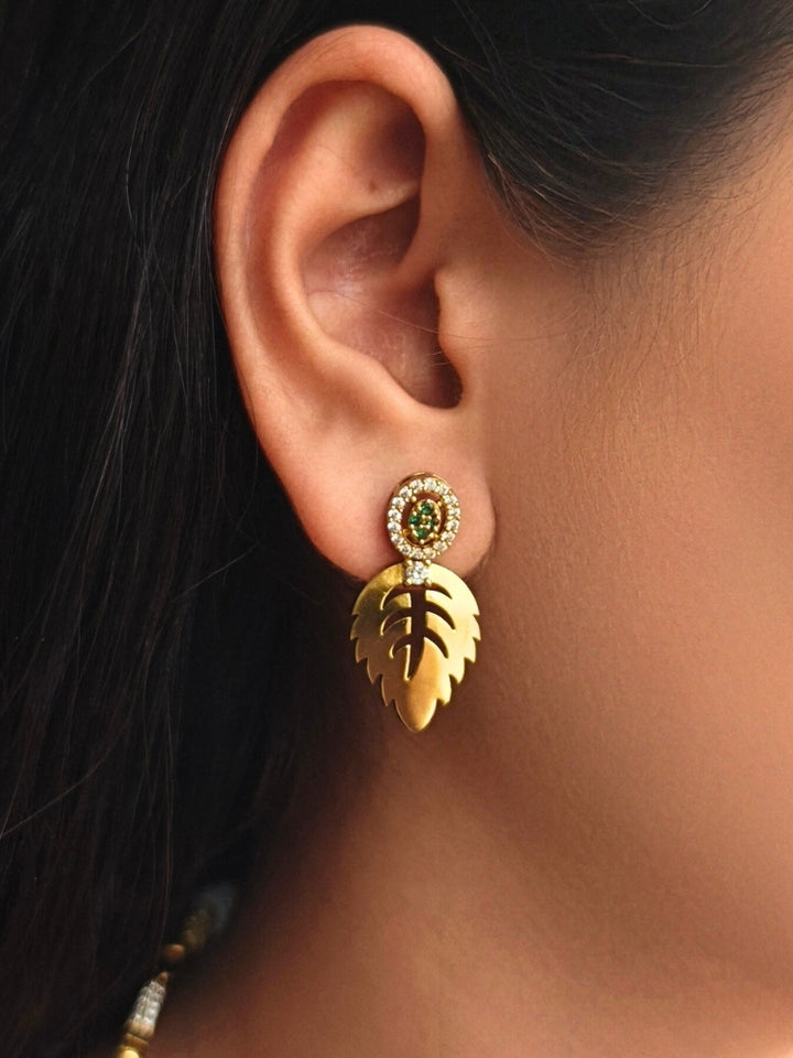 The Maple Green Necklace Set, featuring gold and green tones with matching leaf-shaped pendants and sparkling American Diamonds, is displayed on a black surface with flowers—ideal for wedding ceremony jewelry.