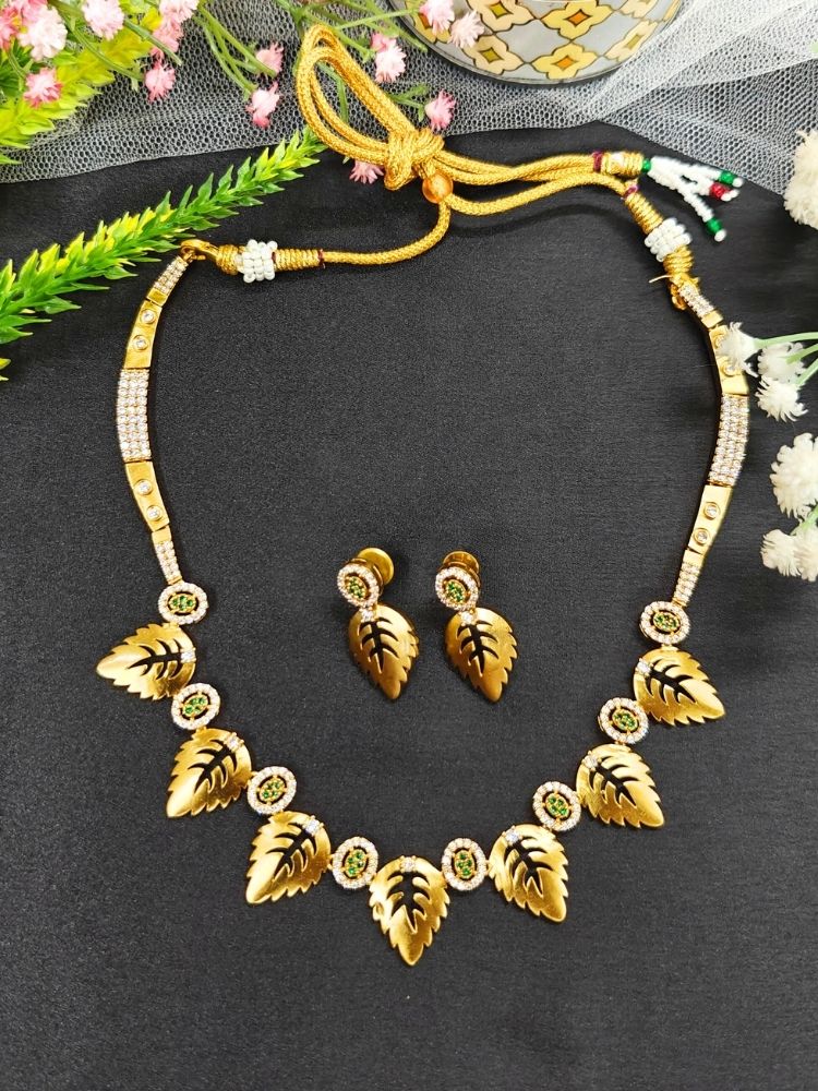 The Maple Green Necklace Set, featuring gold and green tones with matching leaf-shaped pendants and sparkling American Diamonds, is displayed on a black surface with flowers—ideal for wedding ceremony jewelry.