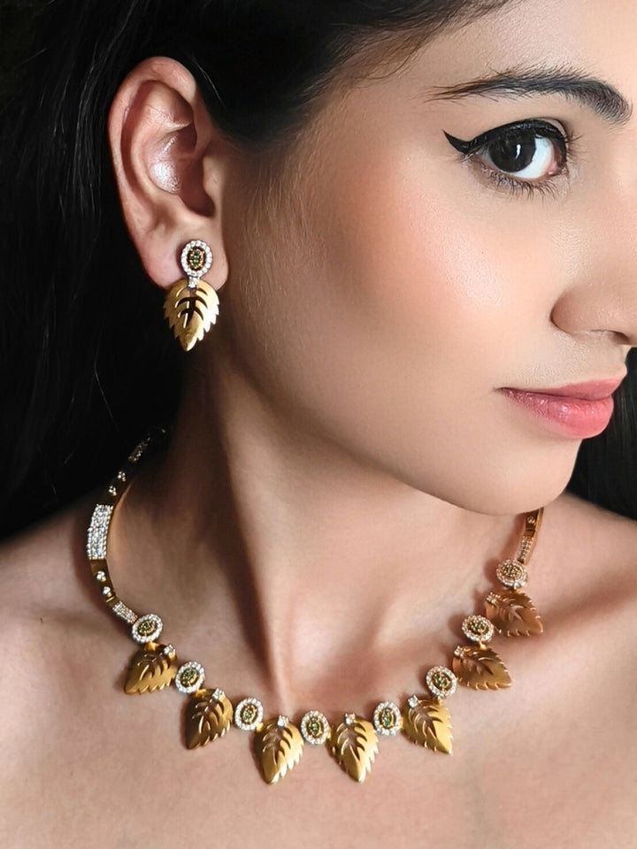The Maple Green Necklace Set, featuring gold and green tones with matching leaf-shaped pendants and sparkling American Diamonds, is displayed on a black surface with flowers—ideal for wedding ceremony jewelry.