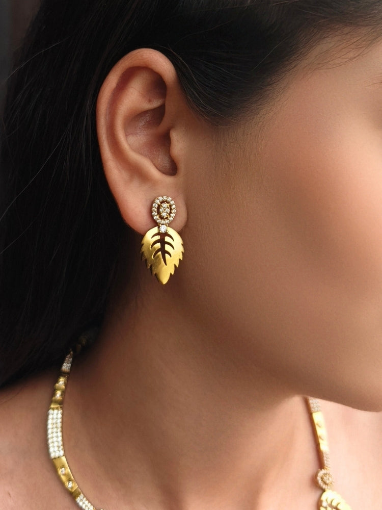 The Maple White Necklace Set, featuring gold and White tones with matching leaf-shaped pendants and sparkling American Diamonds, is displayed on a black surface with flowers—ideal for wedding ceremony jewelry.