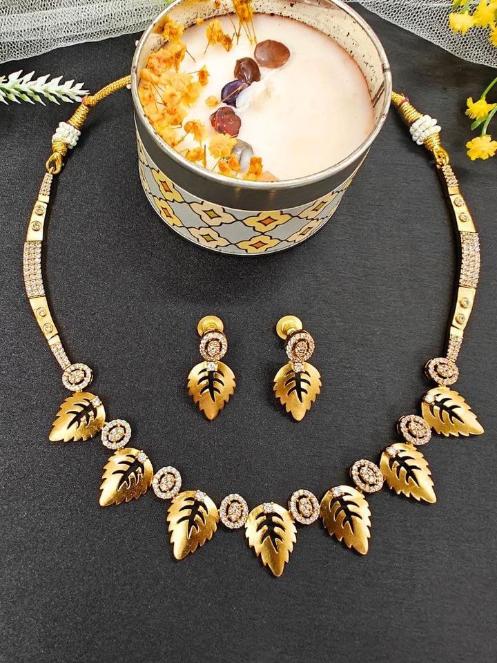 The Maple White Necklace Set, featuring gold and White tones with matching leaf-shaped pendants and sparkling American Diamonds, is displayed on a black surface with flowers—ideal for wedding ceremony jewelry.