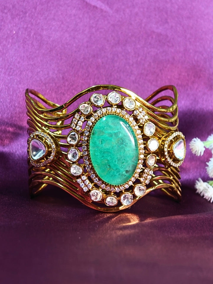 The Mariam Mint Green Bracelet (Adjustable) features an intricate wavy gold design with a large oval mint green stone at the center, accented by small kundan stones, and is displayed on a purple satin backdrop—ideal for elegant wedding ensembles.