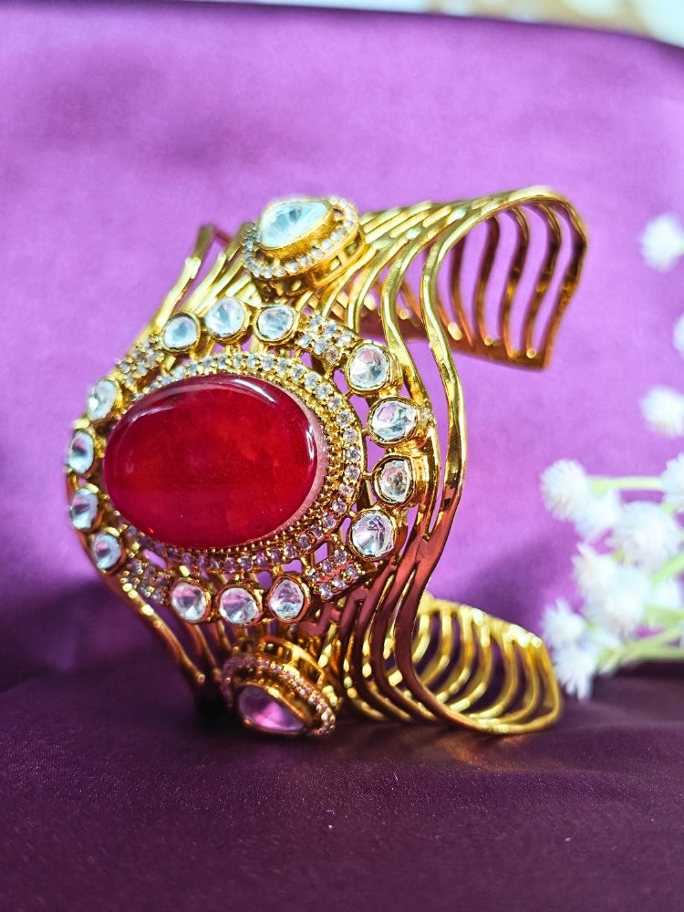 The Mariam Pink Bracelet (Adjustable) features an intricate wavy gold design with a large oval Pink stone at the center, accented by small kundan stones, and is displayed on a purple satin backdrop—ideal for elegant wedding ensembles.
