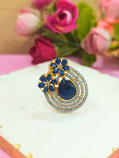 The Meadow Blue Ring is an adjustable Cocktail Ring featuring a large blue gemstone center, American diamonds in three concentric circles, and blue stone flowers, showcased on a white surface with pink roses in the background.