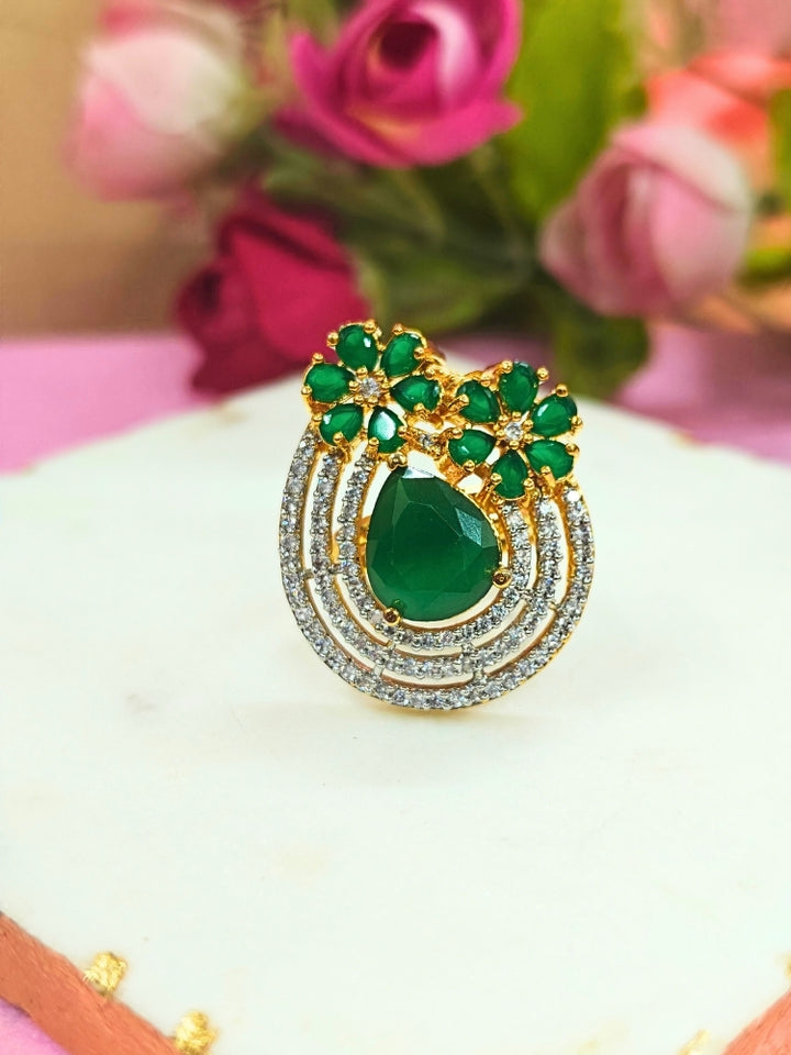 The Meadow Green Ring is an adjustable Cocktail Ring featuring a large Green gemstone center, American diamonds in three concentric circles, and Green stone flowers, showcased on a white surface with pink roses in the background.