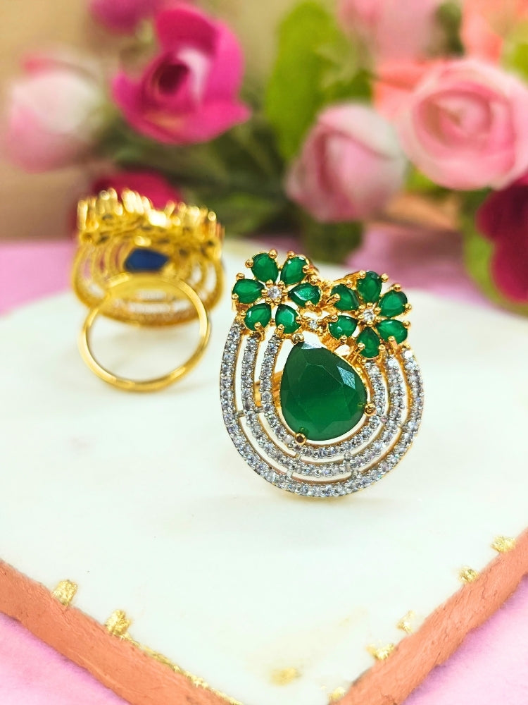 The Meadow Green Ring is an adjustable Cocktail Ring featuring a large Green gemstone center, American diamonds in three concentric circles, and Green stone flowers, showcased on a white surface with pink roses in the background.