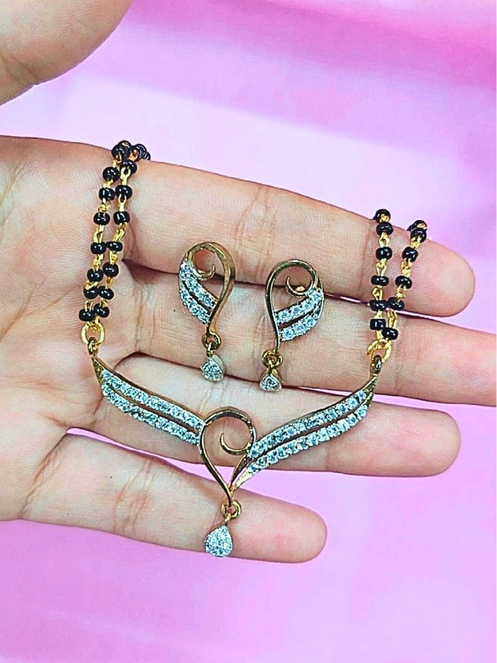 A hand holds the Meenakshi Mangalsutra—a gold necklace with black mangalsutra beads and matching earrings, pendant featuring sparkling American Diamonds—set against a soft pink background, perfect for your wedding ceremony dress. Diamond mangalsutra. Modern mangalsutra. 