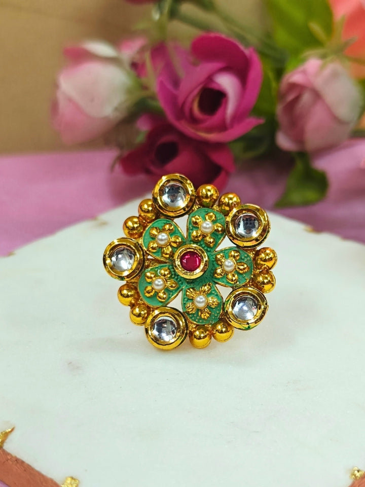 The Meenu Green Ring, a gold adjustable piece with floral patterns and embedded clear gemstones, is shown on a white surface with pink roses in the blurred background—perfect for your wedding ceremony dress.