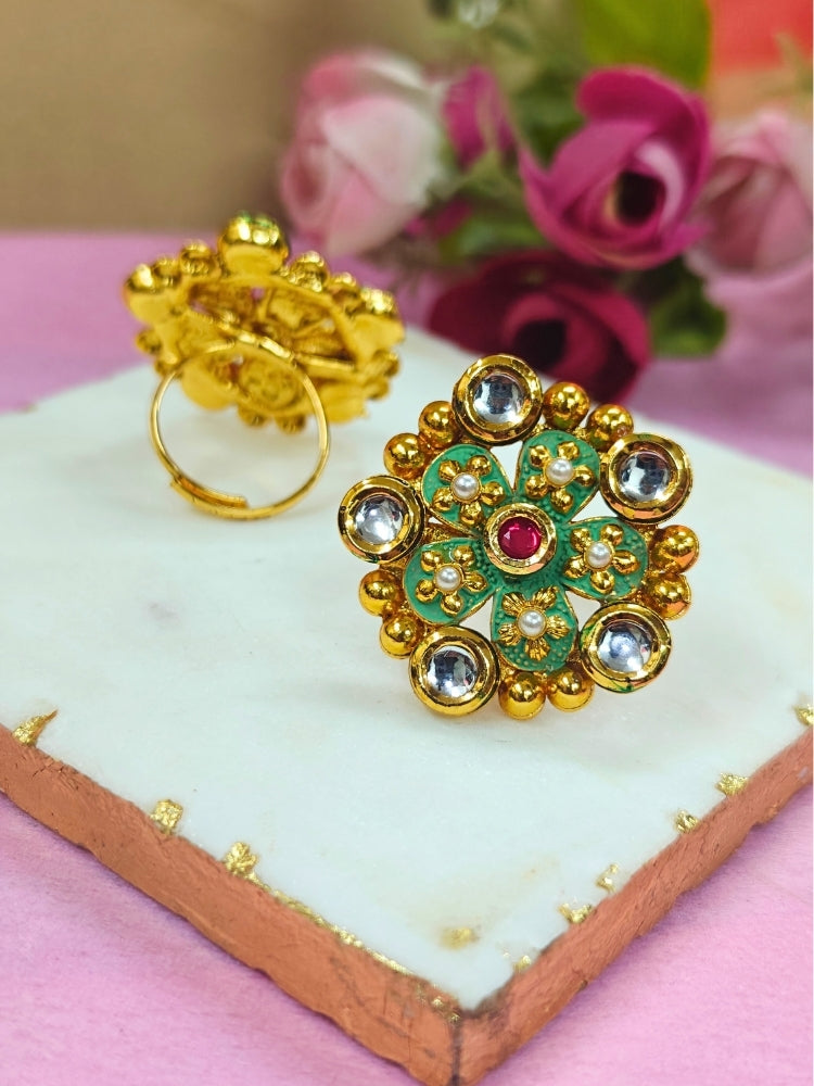 The Meenu Green Ring, a gold adjustable piece with floral patterns and embedded clear gemstones, is shown on a white surface with pink roses in the blurred background—perfect for your wedding ceremony dress.