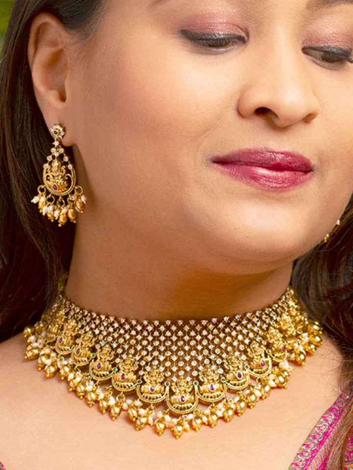 A woman models the Meherin Choker Necklace Set—an elegant gold choker necklace set with jali work along with american diamonds and Goddess Laxmi circumfrencing the bottom, along with matching earrings —paired with a pink outfit. Only her lower face is shown, emphasizing the set’s intricate design.