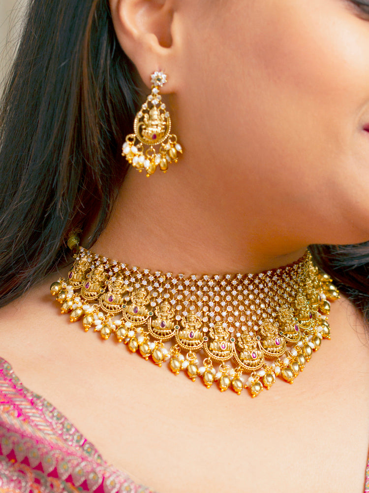 A woman models the Meherin Choker Necklace Set—an elegant gold choker necklace set with jali work along with american diamonds and Goddess Laxmi circumfrencing the bottom, along with matching earrings —paired with a pink outfit. Only her lower face is shown, emphasizing the set’s intricate design.