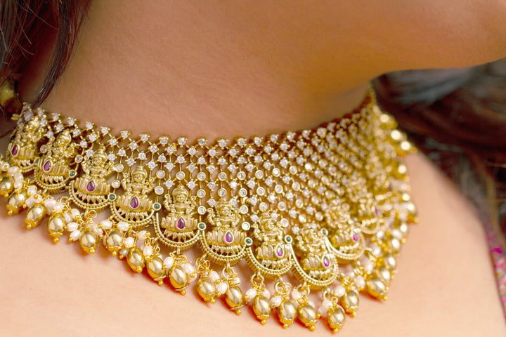 A woman models the Meherin Choker Necklace Set—an elegant gold choker necklace set with jali work along with american diamonds and Goddess Laxmi circumfrencing the bottom, along with matching earrings —paired with a pink outfit. Only her lower face is shown, emphasizing the set’s intricate design.
