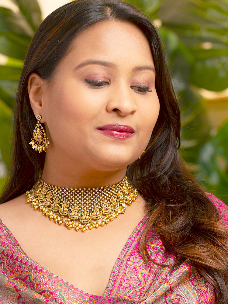 A woman models the Meherin Choker Necklace Set—an elegant gold choker necklace set with jali work along with american diamonds and Goddess Laxmi circumfrencing the bottom, along with matching earrings —paired with a pink outfit. Only her lower face is shown, emphasizing the set’s intricate design.