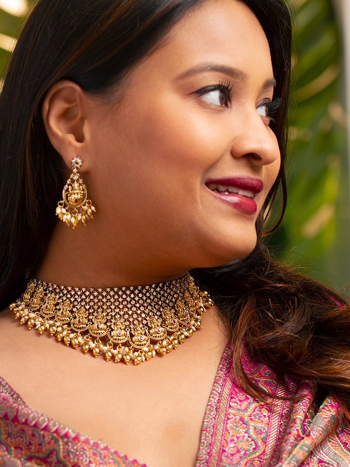A woman models the Meherin Choker Necklace Set—an elegant gold choker necklace set with jali work along with american diamonds and Goddess Laxmi circumfrencing the bottom, along with matching earrings —paired with a pink outfit. Only her lower face is shown, emphasizing the set’s intricate design.