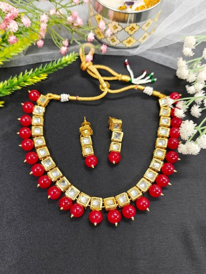 The Mehul Cherry Red Necklace Set, featuring gold and white Kundan stones with large cherry red beads and matching square earrings, is displayed on a black background with white and pink flowers and a decorative container.