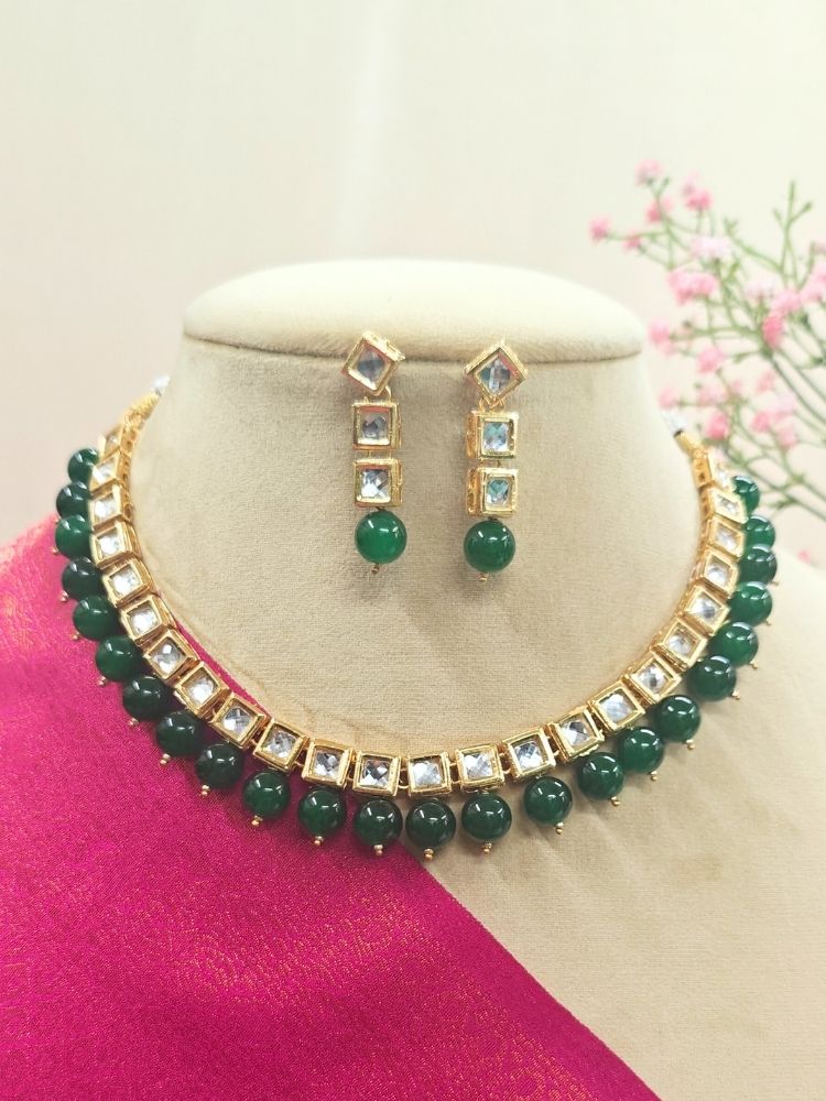 The Green Necklace Set, featuring gold and white Kundan stones with large Green beads and matching square earrings, is displayed on a cream dummy with pink cloth. 