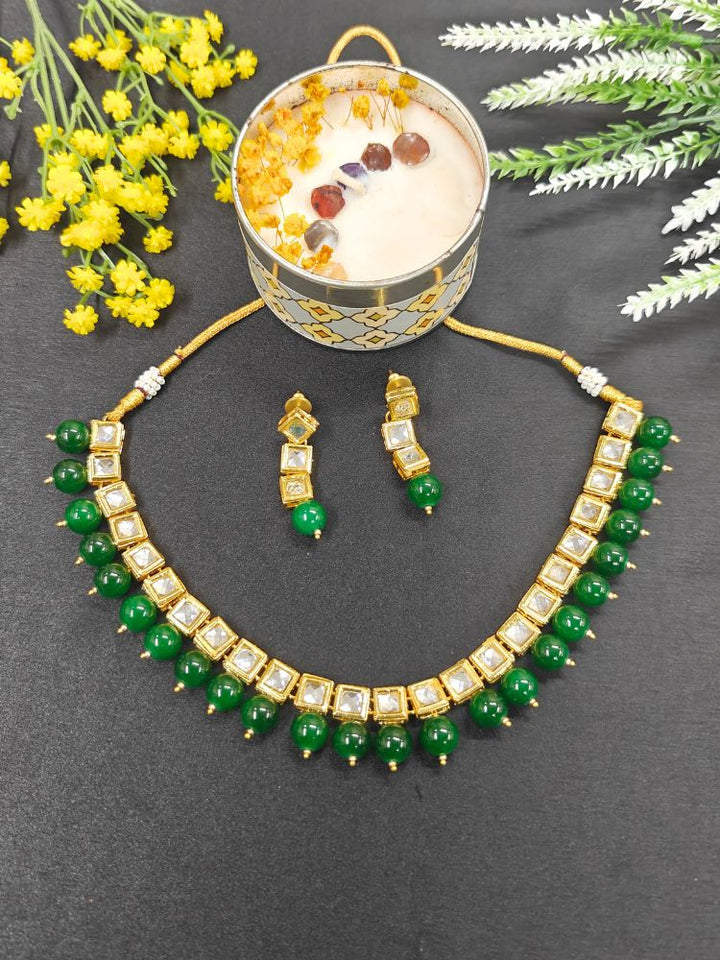 The Green Necklace Set, featuring gold and white Kundan stones with large Green beads and matching square earrings, is displayed on a black background with white and pink flowers and a decorative container.