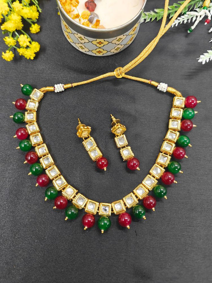 The Mehul Cherry Red & Green Necklace Set, featuring gold and white Kundan stones with large cherry red & Green beads and matching square earrings, is displayed on a black background with white and pink flowers and a decorative container.