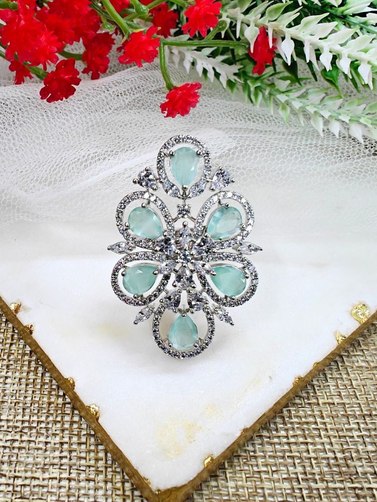 The Melissa Mint Green cocktail Ring is a sparkling silver ring with an intricate floral design, featuring Mint Green teardrop gemstones and clear American Diamonds, elegantly displayed on a white marble slab with red and white flowers in the background.