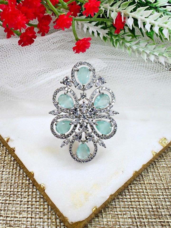 The Melissa Mint Green cocktail Ring is a sparkling silver ring with an intricate floral design, featuring Mint Green teardrop gemstones and clear American Diamonds, elegantly displayed on a white marble slab with red and white flowers in the background.