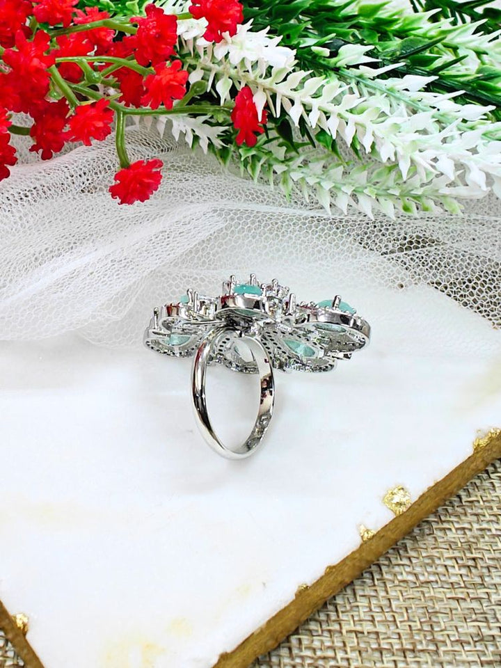 The Melissa Mint Green cocktail Ring is a sparkling silver ring with an intricate floral design, featuring Mint Green teardrop gemstones and clear American Diamonds, elegantly displayed on a white marble slab with red and white flowers in the background.